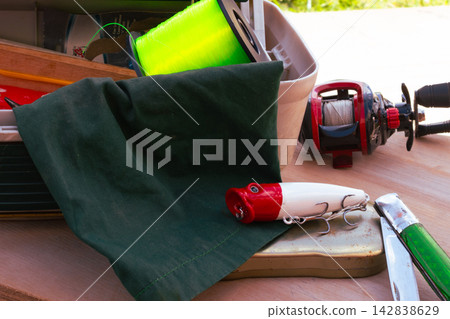 Fishing equipment on a wooden surface. Including a fishing reel with line. A bright green spool of fishing line. A red and white fishing lure. And a pocket knife Fishing equipment on a wooden surface. Including a fishing reel with line. A bright green spool of fishing line. A red and white fishing lure. And a pocket knife 142838629