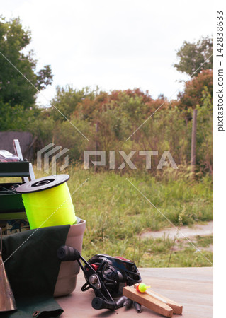 Fishing tackle arranged on a wooden surface with a bright yellow line spool, black baitcasting reel, bobbers and weights against a soft green outdoor background, ready for angling Fishing tackle arranged on a wooden surface with a bright yellow line spool, black baitcasting reel, bobbers and weights against a soft green outdoor background, ready for angling 142838633
