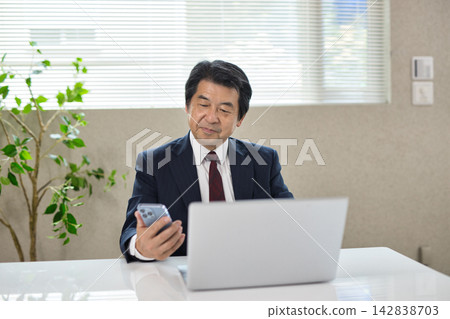 A senior man holding a smartphone while looking at a computer. A senior man holding a smartphone while looking at a computer. 142838703