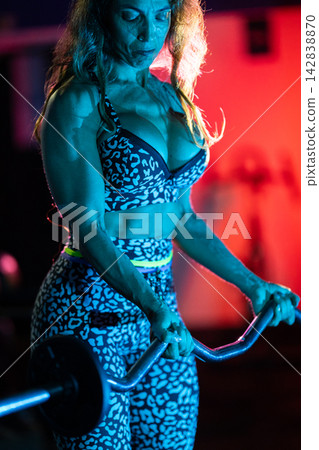 Woman lifts barbell in gym during night workout session with colorful lighting and focused expression Woman lifts barbell in gym during night workout session with colorful lighting and focused expression 142838870