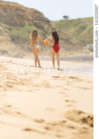 Two women walking on sandy beach holding an orange flotation device, wearing swimsuits, with coastal cliffs and ocean waves in the background under a clear sky Two women walking on sandy beach holding an orange flotation device, wearing swimsuits, with coastal cliffs and ocean waves in the background under a clear sky 142838987