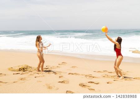 Two women playing with a beach ball on sandy shore, ocean waves crashing in the background under a clear sky during a sunny day at the beach Two women playing with a beach ball on sandy shore, ocean waves crashing in the background under a clear sky during a sunny day at the beach 142838988