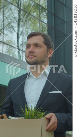 Vertical video. Businessman in despair lost job and walking near office center with carton box and documents with a table flower. Unemployment rate growing due pandemic. Vertical video. Businessman in despair lost job and walking near office center with carton box and documents with a table flower. Unemployment rate growing due pandemic. 142839210