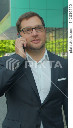 Vertical video. Caucasian businessman in glasses walking the street at the office center and talking on the phone. Close up. Rear. Outside. Vertical video. Caucasian businessman in glasses walking the street at the office center and talking on the phone. Close up. Rear. Outside. 142839220