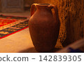 Old clay red pot with two handles standing near wall of samand or plaster against background of blurred background of dark room, illuminated by orange light, on floor carpet with national patterns 142839305