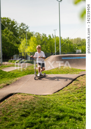 Fair-haired teen riding a scooter in the park on a greenery background 142839404