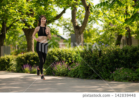 Slim young woman in black sportswear and headphones running in a park Slim young woman in black sportswear and headphones running in a park 142839448