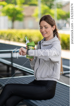 Young woman in grey sweatshirt with a green bottle in hand looking at her watch Young woman in grey sweatshirt with a green bottle in hand looking at her watch 142839474