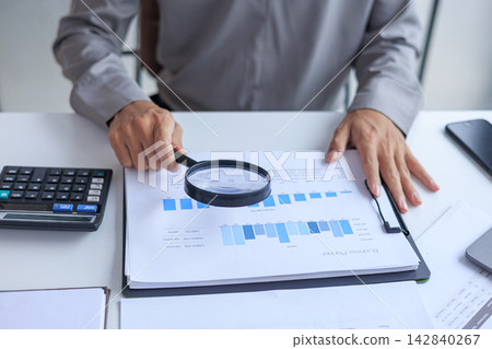 Person using a magnifying glass to review financial and business growth bar charts on a white desk. Person using a magnifying glass to review financial and business growth bar charts on a white desk. 142840267