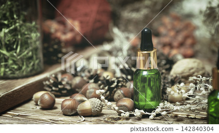 A green glass bottle of natural essential oil among acorns, pinecones and dry herbs on wooden surface, authentic organic extract, selected focus. A green glass bottle of natural essential oil among acorns, pinecones and dry herbs on wooden surface, authentic organic extract, selected focus. 142840304
