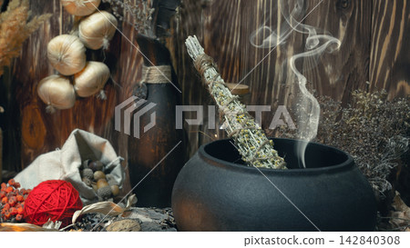 Pagan witch magic altar. Smoking twist of bitter wormwood herb for fumigation in cast iron cauldron, ritual objects in wooden background, dark key, selective focus. Pagan witch magic altar. Smoking twist of bitter wormwood herb for fumigation in cast iron cauldron, ritual objects in wooden background, dark key, selective focus. 142840308