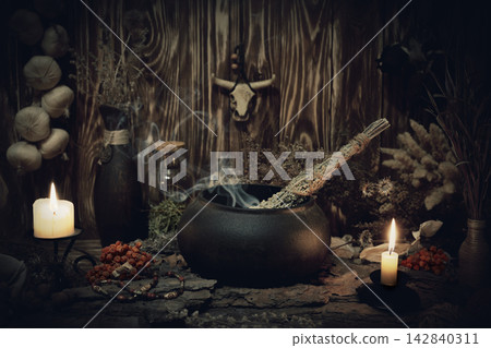 Authentic forest witch altar. Burning candles, dry herbs, iron cauldron, smoking mugwort, bulls skull mojo, bunch of garlic in wooden background in the dark, low key, selective focus. Authentic forest witch altar. Burning candles, dry herbs, iron cauldron, smoking mugwort, bulls skull mojo, bunch of garlic in wooden background in the dark, low key, selective focus. 142840311