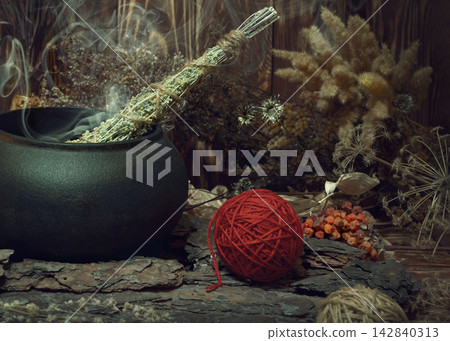 Magic items. Smoking twist of bitter wormwood herb for fumigation in iron cauldron, a ball of red wool, pine bark and dry flowers on wooden background, dark key, selective focus. 142840313