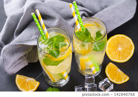 Cold homemade lemonade with fresh lemon, mint and ice in glasses on black textured stone background Cold homemade lemonade with fresh lemon, mint and ice in glasses on black textured stone background 142840788