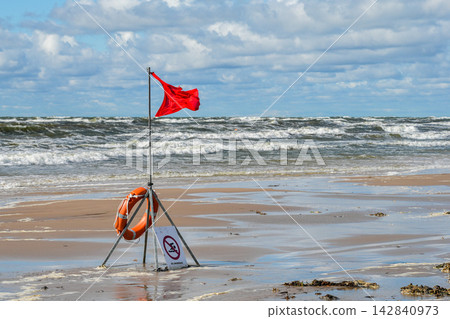 Red Warning Flag and Lifebuoy on Windy Baltic Sea Beach with Rough Waves and Wet Sandy Shore 142840973