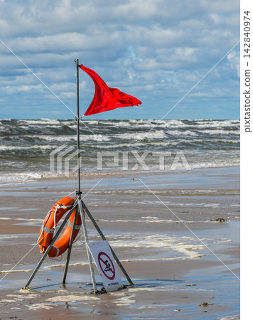 Red Warning Flag and Lifebuoy on Stormy Baltic Sea Beach with Rough Waves and Cloudy Sky Red Warning Flag and Lifebuoy on Stormy Baltic Sea Beach with Rough Waves and Cloudy Sky 142840974