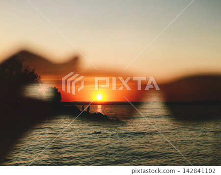 Dreamy sunset over calm water with two silhouetted people on a rocky shore, glowing reflections and blurred foreground creating a nostalgic atmospheric summer landscape for travel and lifestyle image Dreamy sunset over calm water with two silhouetted people on a rocky shore, glowing reflections and blurred foreground creating a nostalgic atmospheric summer landscape for travel and lifestyle image 142841102