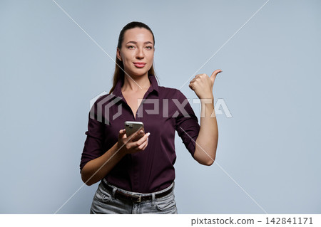 Young businesswoman holding smartphone and pointing thumb to side against grey background Young businesswoman holding smartphone and pointing thumb to side against grey background 142841171