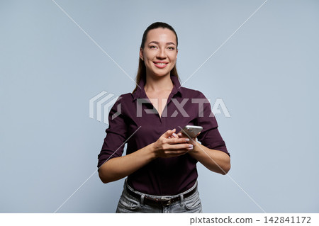 Young professional woman holding smartphone smiling at camera on light background Young professional woman holding smartphone smiling at camera on light background 142841172