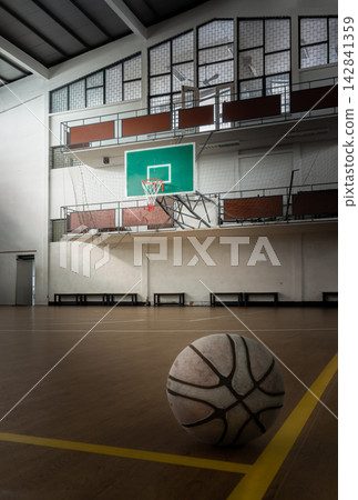 A weathered basketball rests on the court floor in a low-angle perspective, capturing a subtle interplay of light and shadow. Diffused natural light illuminates the ball's worn texture, set against 142841359
