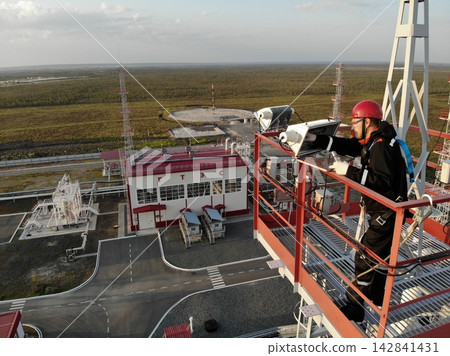 Man in safety helmet examines instruments from tall steel platform at gas installation, alert and focused. Man stands above roads, pipes, and service buildings, with wide marshland stretching beyond Man in safety helmet examines instruments from tall steel platform at gas installation, alert and focused. Man stands above roads, pipes, and service buildings, with wide marshland stretching beyond 142841431