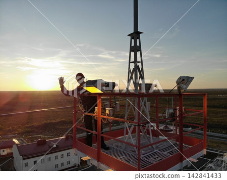 Worker in helmet stands beside rooftop equipment and raises one hand on elevated platform. Warm sunset light silhouettes industrial hardware above facility buildings and open terrain. Worker in helmet stands beside rooftop equipment and raises one hand on elevated platform. Warm sunset light silhouettes industrial hardware above facility buildings and open terrain. 142841433