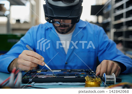 Technician man soldering technician dressed in a blue lab coat is diligently repairing a laptop Technician man soldering technician dressed in a blue lab coat is diligently repairing a laptop 142842068