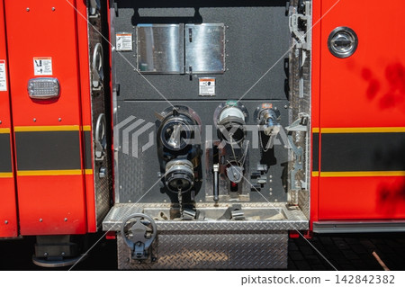 A close detail shows the pump panel and hose connections on a red fire truck. The valves, gauges, metal controls, warning labels, and emergency equipment are shown in bright sunlight. A close detail shows the pump panel and hose connections on a red fire truck. The valves, gauges, metal controls, warning labels, and emergency equipment are shown in bright sunlight. 142842382