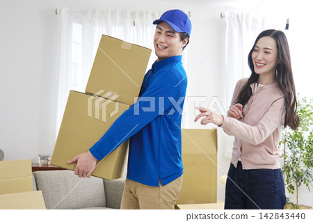 A moving company worker holding cardboard boxes and a woman giving instructions with a smile. A moving company worker holding cardboard boxes and a woman giving instructions with a smile. 142843440