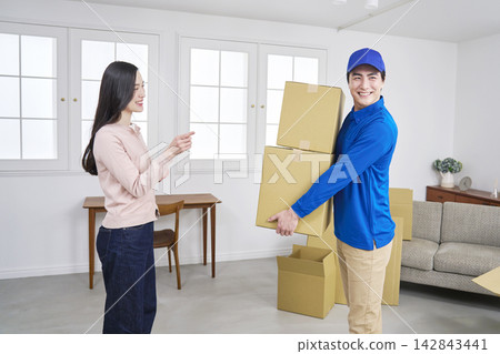 A moving company worker holding cardboard boxes and a woman giving instructions with a smile. A moving company worker holding cardboard boxes and a woman giving instructions with a smile. 142843441