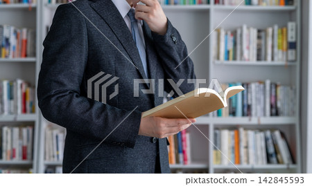 一個穿著西裝的男人正在書架前讀書,陷入沉思。 一個穿著西裝的男人正在書架前讀書,陷入沉思。 142845593