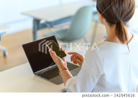 一位女性在辦公室工作,使用筆記型電腦和智慧型手機。 一位女性在辦公室工作,使用筆記型電腦和智慧型手機。 142845616