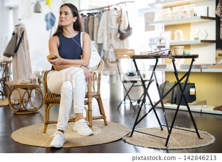 Girl in summer clothes sits on wicker chair in clothing store and looks at fashionable gifts and clothes Girl in summer clothes sits on wicker chair in clothing store and looks at fashionable gifts and clothes 142846213