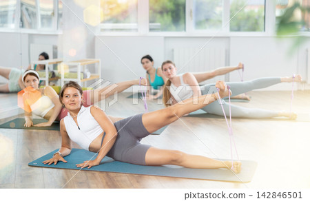 Girls perform exercises lying on side with legs straightened and narrow elastic band on shins Girls perform exercises lying on side with legs straightened and narrow elastic band on shins 142846501