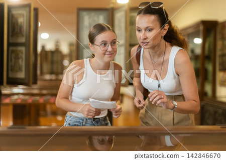 Young woman with teen daughter visitors stands near horizontal glass display case Young woman with teen daughter visitors stands near horizontal glass display case 142846670