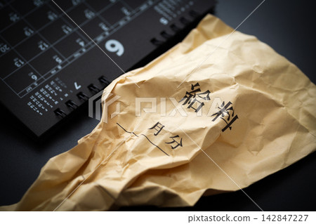 黑色背景上放著一個皺巴巴的工資信封和一張六月的日曆。 黑色背景上放著一個皺巴巴的工資信封和一張六月的日曆。 142847227