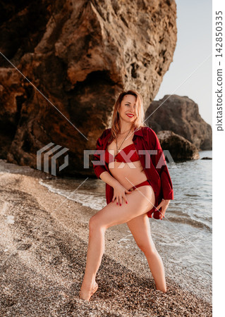 Woman beach bikini posing on a scenic pebble shoreline near large sea rocks at sunset Woman beach bikini posing on a scenic pebble shoreline near large sea rocks at sunset 142850335