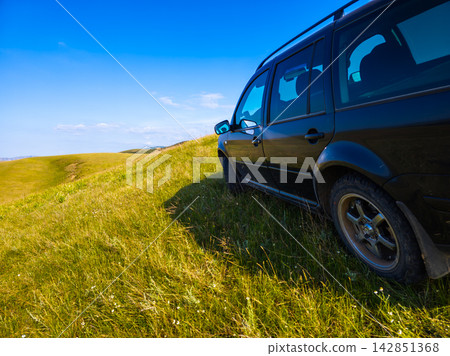Dark Station Wagon on Grassy Hillside Under Blue Sky Dark Station Wagon on Grassy Hillside Under Blue Sky 142851368