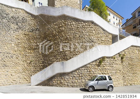 A silver microcar is parked at the base of a large, textured stone wall with a modern, winding white staircase ascending in San Marino A silver microcar is parked at the base of a large, textured stone wall with a modern, winding white staircase ascending in San Marino 142851934