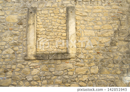 A rough-hewn stone wall features a section of a weathered, stone window frame in the center A rough-hewn stone wall features a section of a weathered, stone window frame in the center 142851943