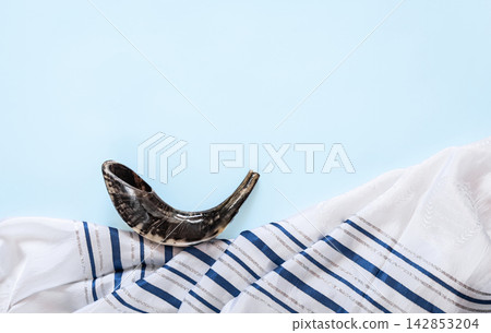 Top view of shofar across tallit on light background, Yom Kippur greeting concept. Jewish holiday banner with tallit and shofar. Copy space for text, nobody Top view of shofar across tallit on light background, Yom Kippur greeting concept. Jewish holiday banner with tallit and shofar. Copy space for text, nobody 142853204
