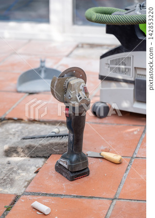 Cordless angle grinder stands beside a construction vacuum cleaner on tiled flooring during renovation work. Cordless angle grinder stands beside a construction vacuum cleaner on tiled flooring during renovation work. 142853220
