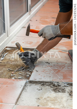 Construction worker breaks and removes old tiles with a hammer and chisel during floor renovation. Construction worker breaks and removes old tiles with a hammer and chisel during floor renovation. 142853241