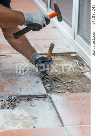 Worker wearing protective gloves removes old ceramic floor tiles using a hammer and chisel during renovation. Worker wearing protective gloves removes old ceramic floor tiles using a hammer and chisel during renovation. 142853242