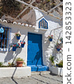 Charming white Mediterranean house with vivid blue door and flower pots nestled beneath rocky cliffs in a picturesque Andalusian coastal settlement 142853323