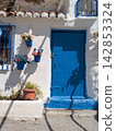 Close up of a vibrant blue wooden door on a whitewashed Andalusian house adorned with colorful flower pots and dramatic sunlit shadows 142853324