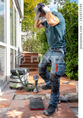 Worker removing ear protection while renovating outdoor patio tiles 142853549