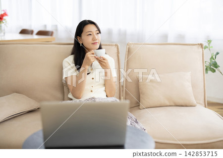 A young Japanese woman relaxes on a sofa by the window, holding a cup of coffee and looking at her laptop. A young Japanese woman relaxes on a sofa by the window, holding a cup of coffee and looking at her laptop. 142853715