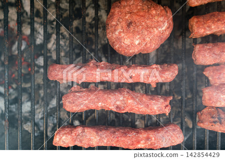Meat patties and sausage rolls cooking on grill over charcoal. Smoky BBQ scene Meat patties and sausage rolls cooking on grill over charcoal. Smoky BBQ scene 142854429