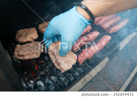 Raw meat patty placed on hot grill with gloved hand. Outdoor BBQ cooking 142854431